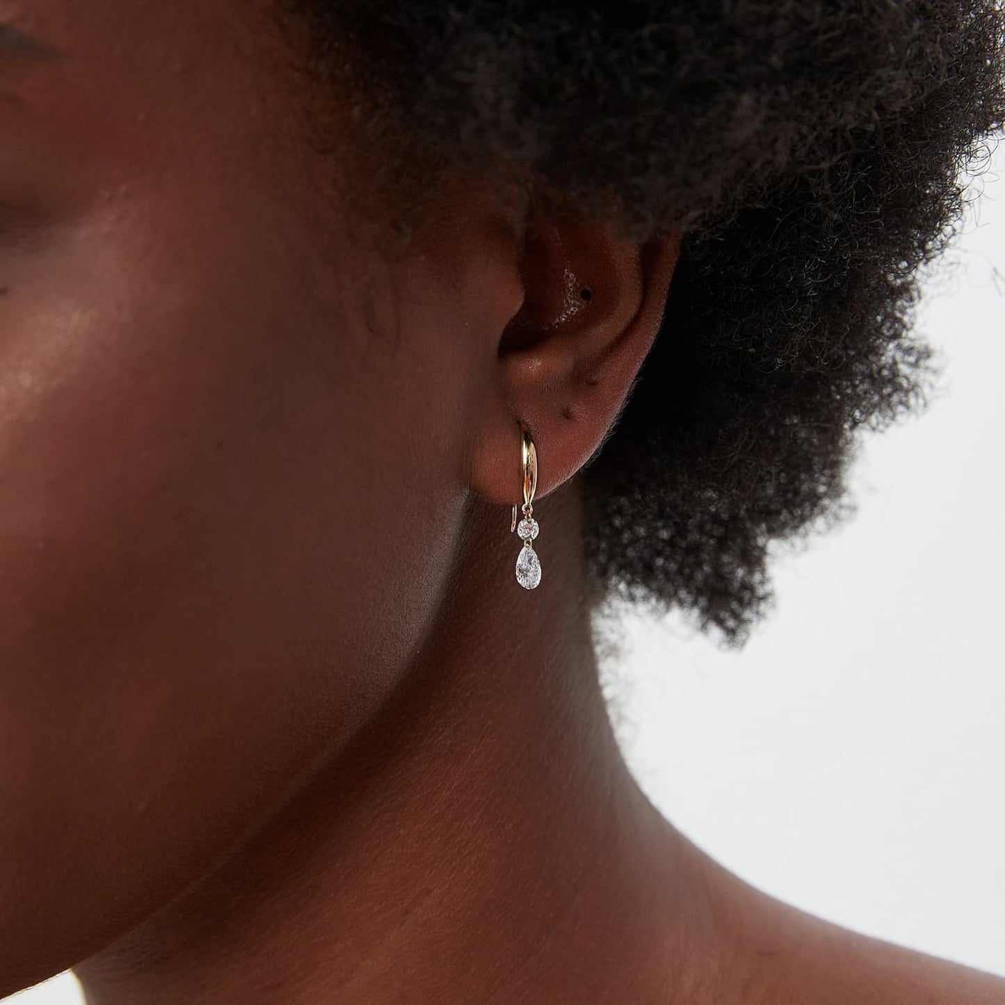 A professional lifestyle photo of the Radiant Drift dangle hoop worn on an ear lobe. Demonstrates the swaying movement of the pear diamond and the dynamic sparkle it brings to a curated 18K gold ear stack.