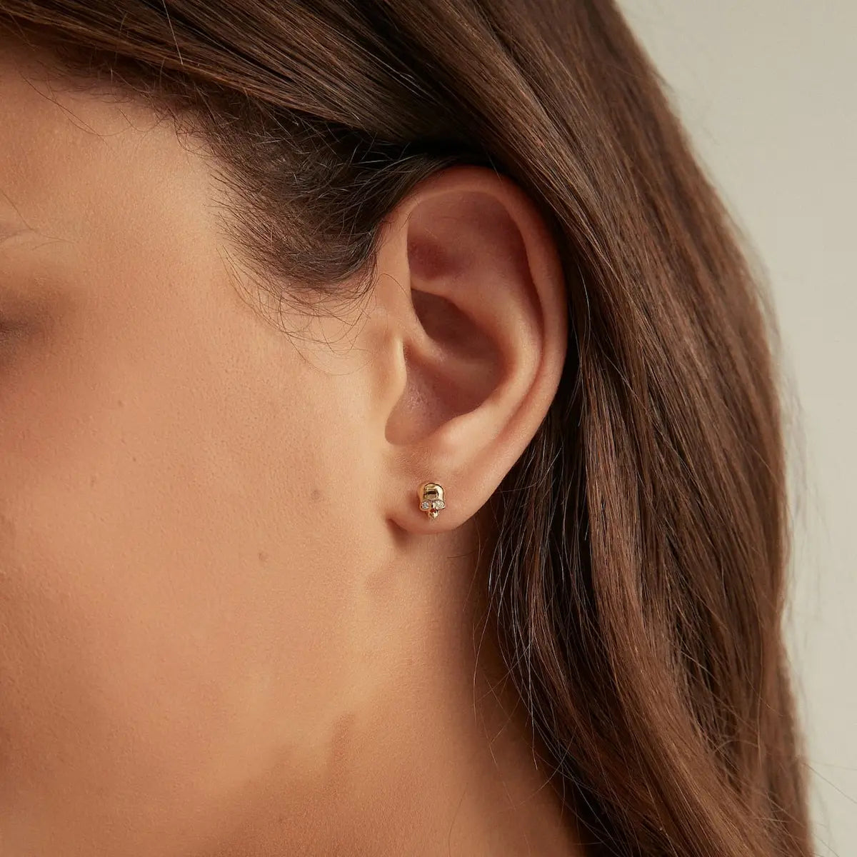 A professional lifestyle photograph showing the Diamond Skull Threaded Stud styled on a model's ear. The image illustrates how the 18K solid gold skull and its brilliant natural diamond catch the light within a curated ear stack. This perspective highlights the edgy, modern silhouette and the compact size of the stud, showcasing its status as a wearable sculpture. The natural lighting emphasizes the fire of the VS1/E diamond, perfect for contemporary luxury jewelry styling inspiration.