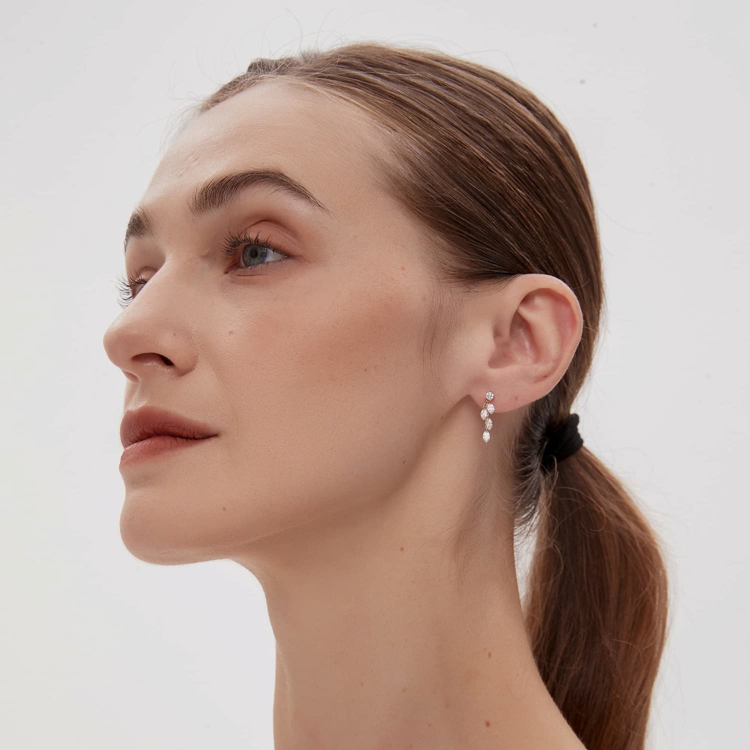 A professional lifestyle photograph showing the Luminous Cascade Dangle Stud styled on a model's ear. The image demonstrates the fluid rhythm of the four cascading marquise diamonds as they follow the movement of the delicate 18K gold chain. This perspective highlights the cinematic radiance of the 0.50ctw diamonds in a real-world setting, illustrating its high-fashion editorial appeal. Captured in natural lighting to emphasize the fire of the D-E color diamonds within a curated ear stack.