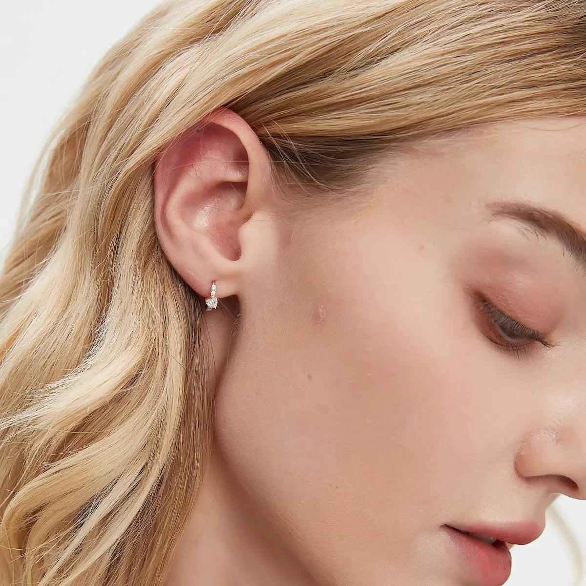 A professional lifestyle photograph showing the Heart Pavé Hoop styled on a model's ear lobe. The 8mm 18K solid gold huggie demonstrates its architectural romance and cinematic radiance within a curated stack. This perspective highlights the modern silhouette of the heart motif and its rhythmic light-catching ability in natural lighting, perfect for a high-fashion, effortless luxury look.