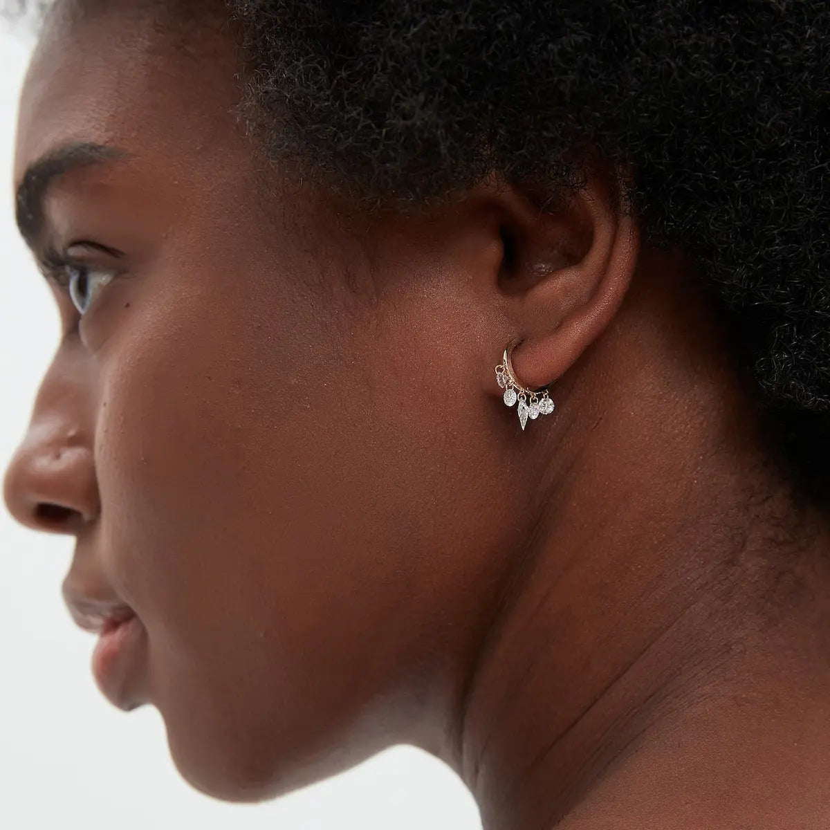 A professional ear stack curation showing the Kite Diamond Chain Dangle Hoop in a lobe piercing. The image demonstrates the vertical movement and fluid motion of the diamond chain against the skin. Perfectly styled with 18K gold studs, this shot illustrates the earring's 8mm inner diameter fit and its ability to add a bold, elongated architectural edge to a layered jewelry look, suitable for daily luxury and sophisticated evening wear.