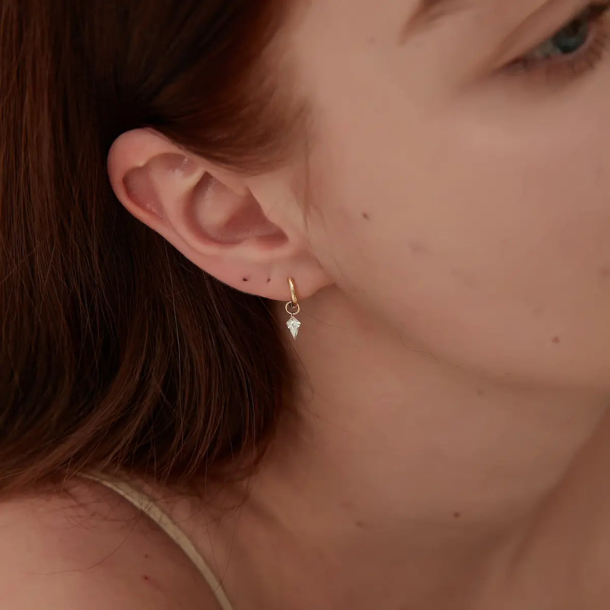 This lifestyle product image demonstrates the dynamic wearability and kinetic elegance of the Kite Diamond Earring Charm from LTM & CO.'s Architectural Rhythm collection, styled delicately on a human ear. The charm is elegantly suspended from a classic 18K solid gold huggie hoop, showcasing its perfect, proportionate scale against the wearer's skin. Thanks to the brand's proprietary laser-drilled floating diamond technology, the kite-shaped lab-grown diamond appears to hover weightlessly. 