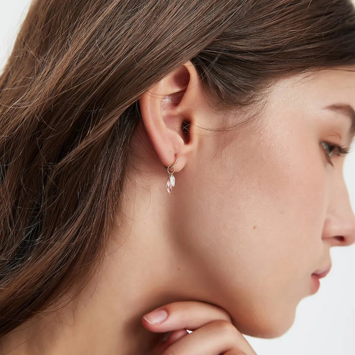 A professional lifestyle photograph showing the Pink Marquise Feather Charm styled on an 18K gold hoop earring. The image illustrates how the marquise diamonds sway gently with movement, adding a soft and romantic accent to a curated ear stack. This perspective highlights the modular design and the fluid, light-catching flutter of the 'Radiant Drift' philosophy. Captured in natural lighting to emphasize the fire of the VVS-VS clarity diamonds and the contrast of the pink stone.