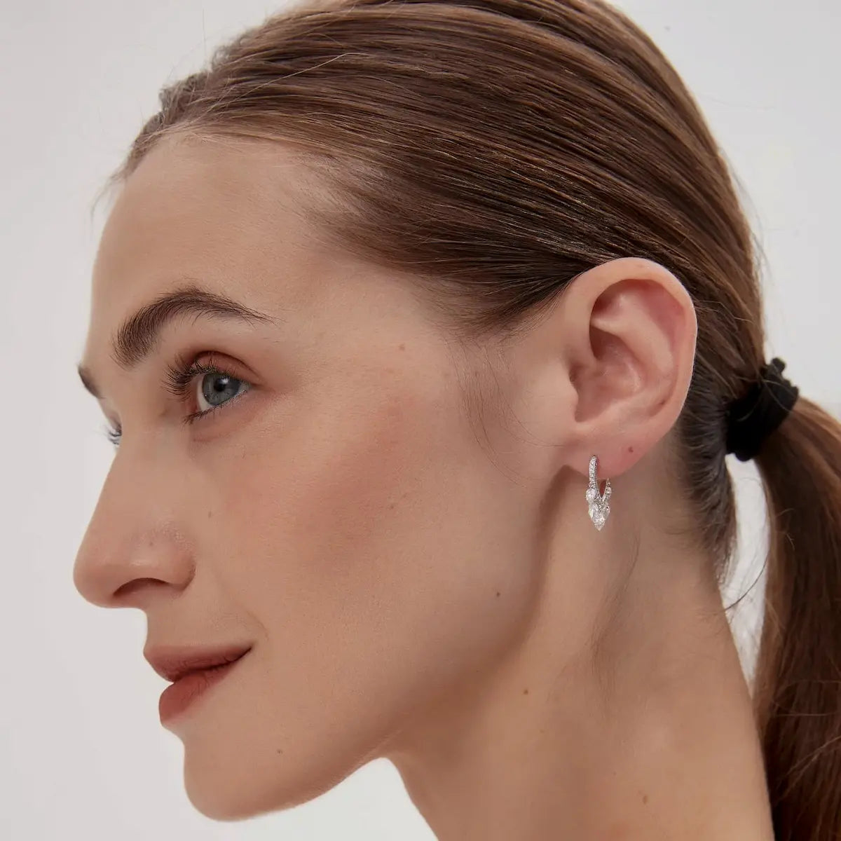 A professional lifestyle image showcasing the Heart Dangle Full Pavé Hoop styled on a model's ear. The piece demonstrates a 360-degree multidimensional glow, capturing light from every angle as the heart-shaped dangle moves. This shot highlights the 2-in-1 versatility of the design, illustrating how the inverted pear diamond and pavé hoop add a sophisticated, romantic narrative to a curated ear stack. Captured in natural lighting to emphasize the intense fire and brilliance of the stones.