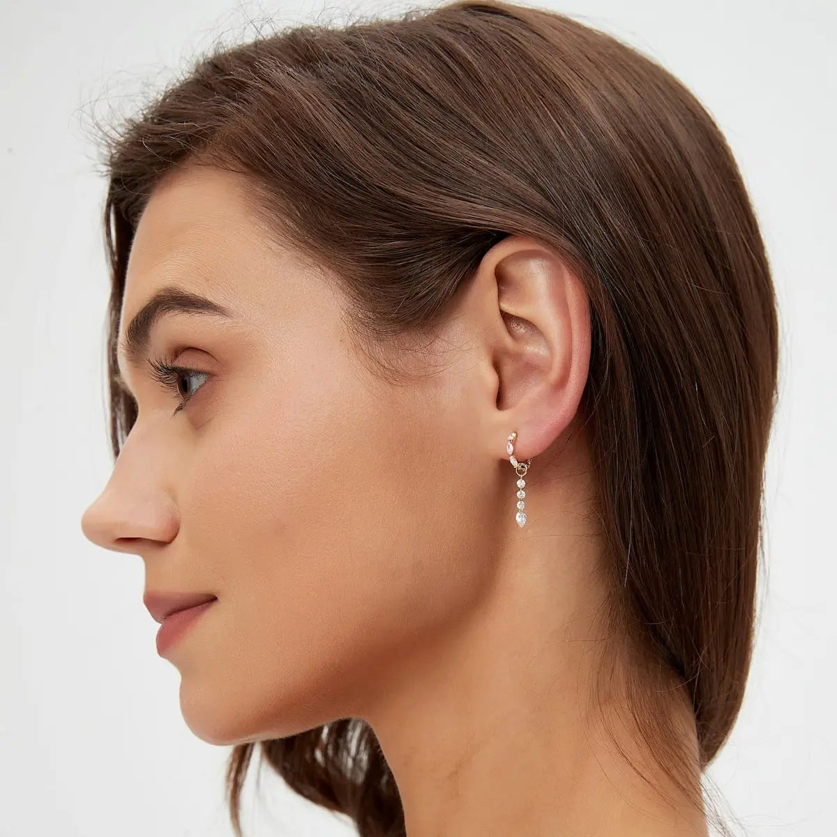 A curated ear stack demonstration featuring the Pear Trio Round Diamond Charm dangling from an 18K gold huggie hoop on a lobe piercing. This styling shot illustrates how the charm adds verticality and a rhythmic glow to a layered earring look. Perfectly paired with minimalist gold studs, the image highlights the charm's scale and its ability to transform basic jewelry into a sophisticated, celestial-themed statement for daily luxury wear.