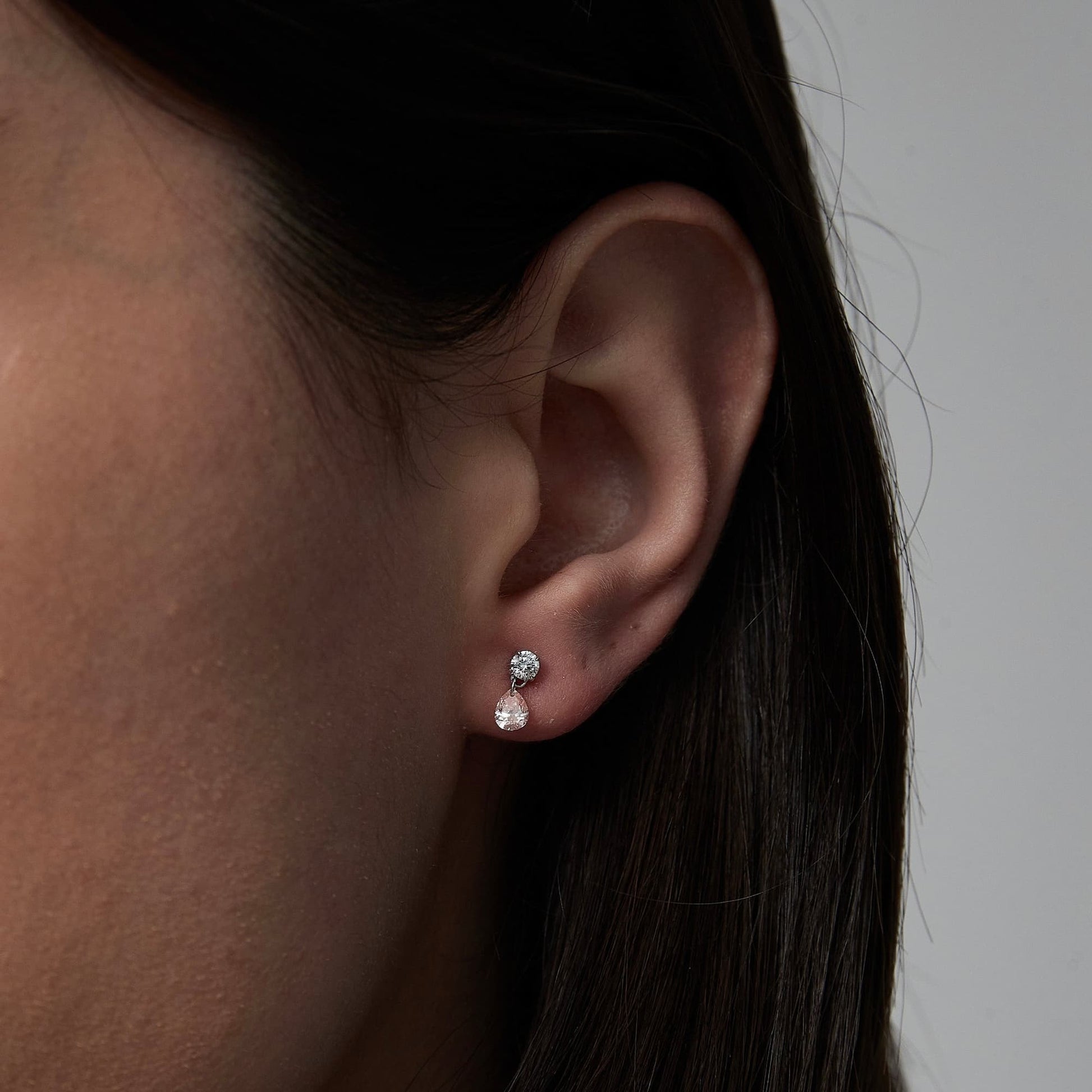 A lifestyle photograph showing the Pink Pear White Round Stud styled on a model's ear. The image demonstrates how the asymmetric pairing of the pink pear and white round diamonds sits perfectly flush against the ear, creating a refined and singular sparkle. This perspective highlights the stud's role in a contemporary curated ear stack, showcasing the vibrant contrast between the two stone shapes. Captured in natural lighting to emphasize the exceptional VVS-VS clarity and fire of the 0.3ctw diamonds.