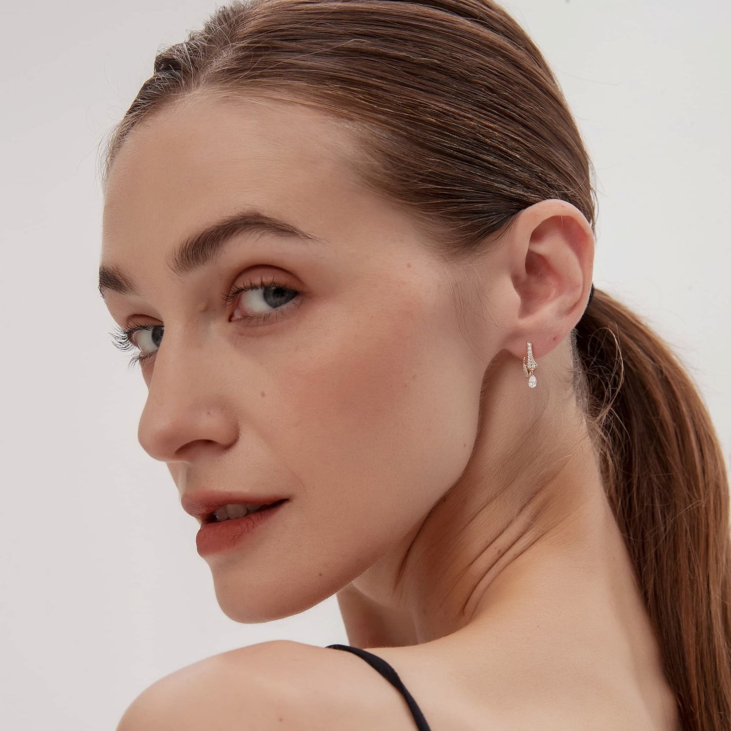 A lifestyle model shot of a woman wearing the Architectural Rhythm collection Serpentine Snake White Diamond Pear Drop Hoop in 18K solid gold. The photograph captures the natural brilliance of the pavé-set body and the fluid movement of the pear-cut diamond dangle as it catches the light. This luxury jewelry piece by LTM Jewels embodies sinuous allure and modern elegance, making it an ideal gift for an anniversary or a bold addition to a sophisticated woman’s fashion collection.